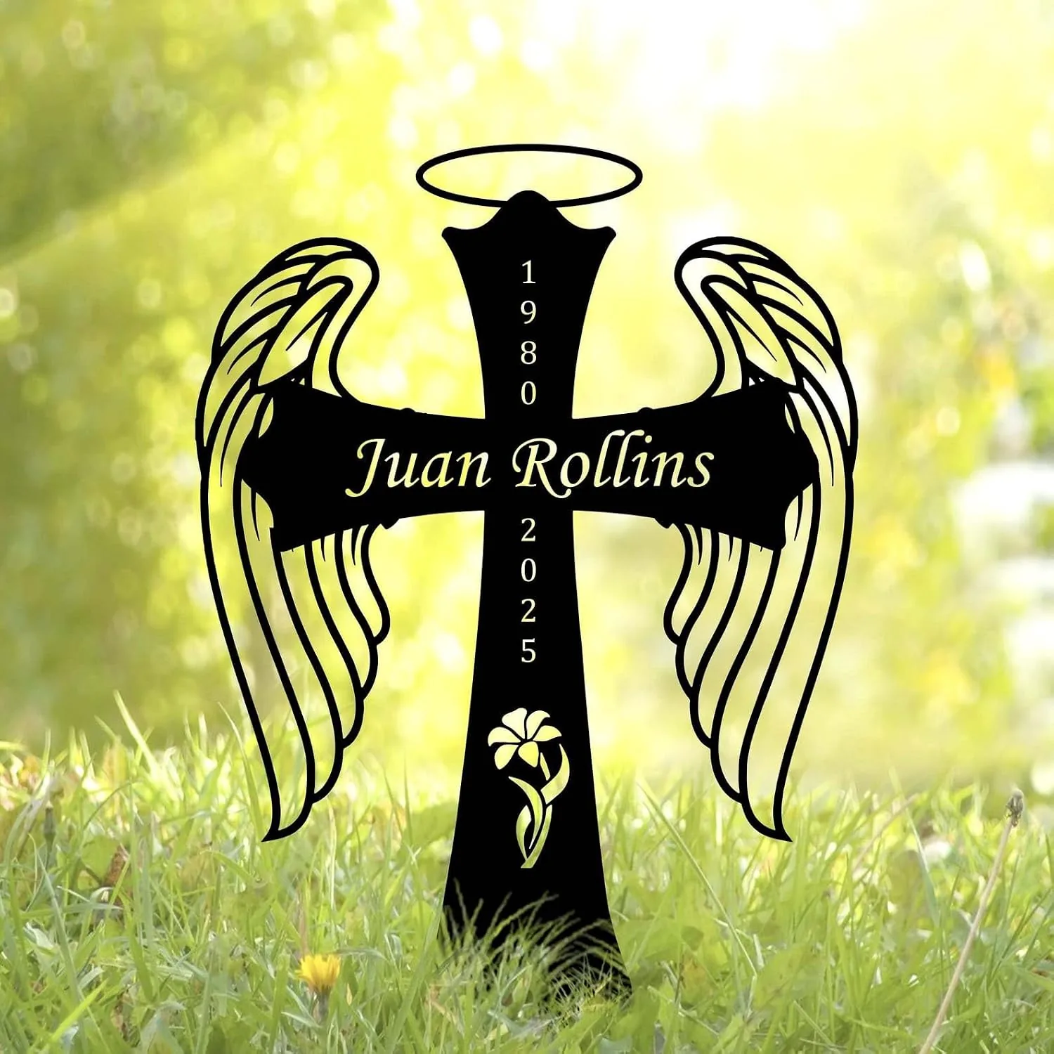 Black angel wing cross yard sign with halo, name "Juan Rollins" across the cross and years 1980–2025, placed in grassy outdoor setting