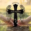 Black angel wing cross memorial stake reading "Juan Rollins" with dates 1980–2025 held by two hands in soil outdoors at sunset