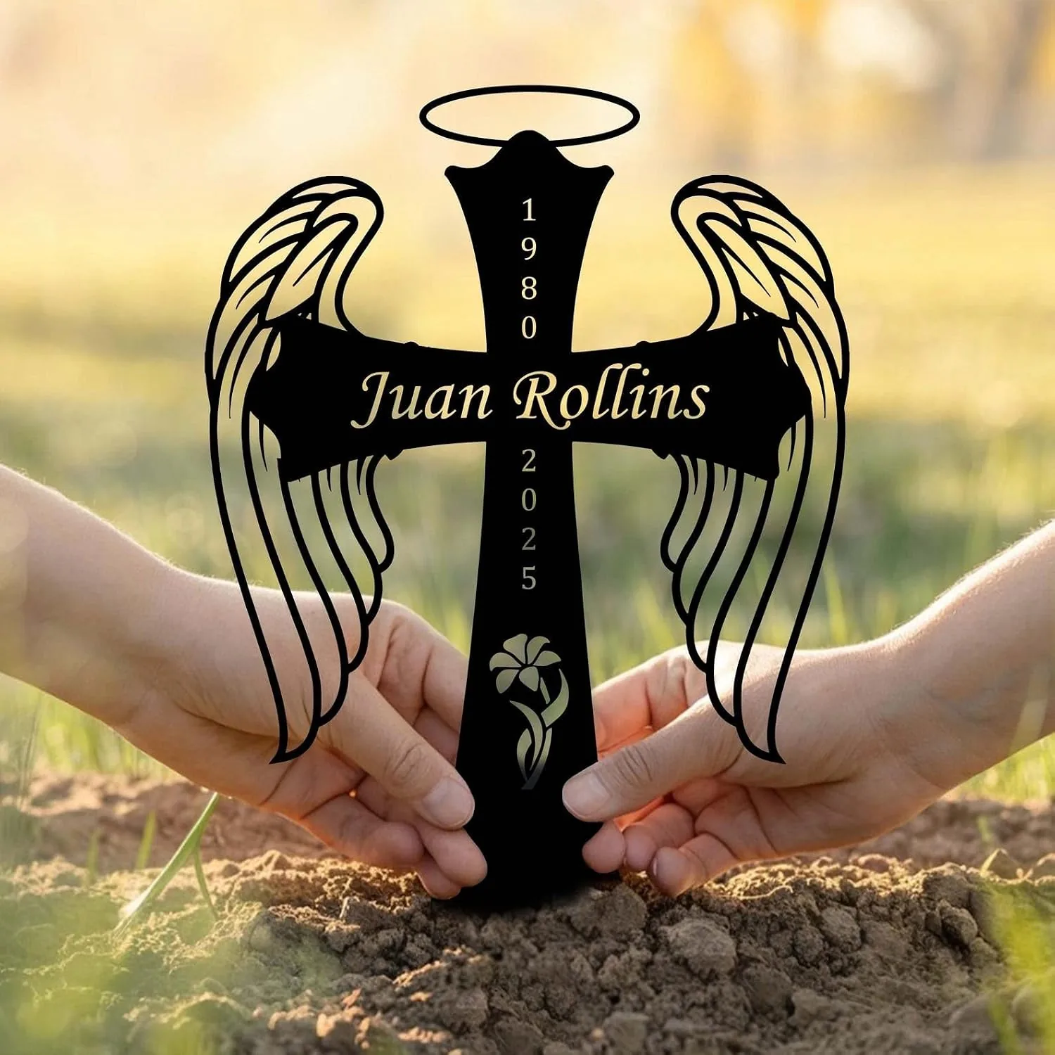 Black angel wing cross memorial stake reading "Juan Rollins" with dates 1980–2025 held by two hands in soil outdoors at sunset