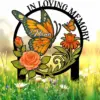 Memorial garden stake with orange butterflies, roses and flowers in a circular black frame reading "In Loving Memory" and the name "Nana" against a sunlit grassy background.
