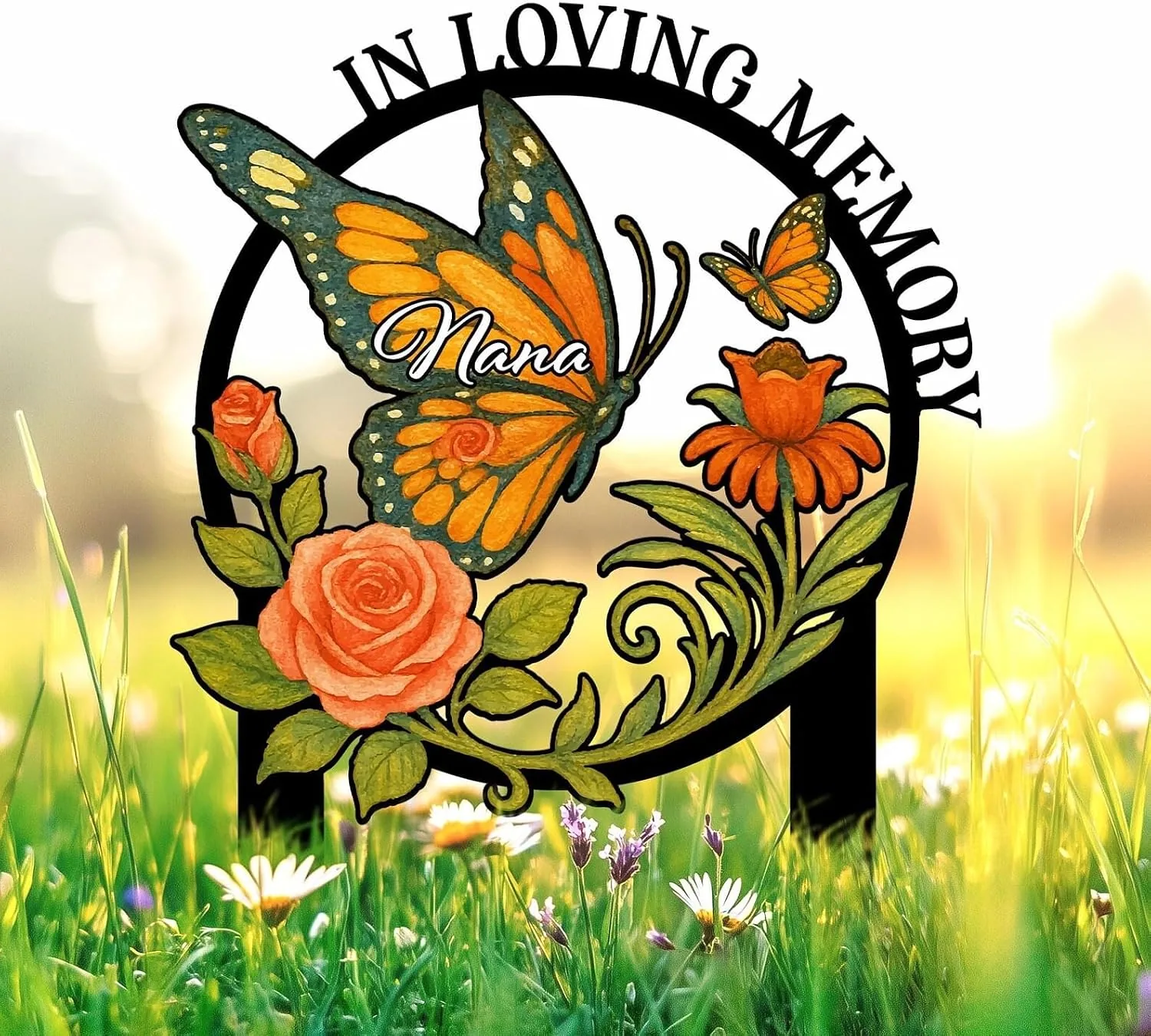Memorial garden stake with orange butterflies, roses and flowers in a circular black frame reading "In Loving Memory" and the name "Nana" against a sunlit grassy background.