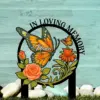 Outdoor memorial sign with painted orange monarch butterfly, roses and flowers on a turquoise wood background, text reads "In Loving Memory" and "Nana"