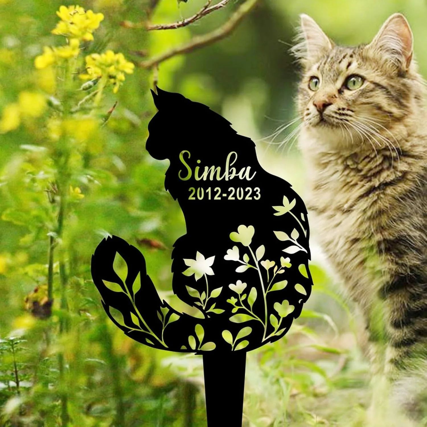 Black metal cat silhouette garden stake reading "Simba 2012-2023" with floral cutouts set among green plants and a tabby cat in the background