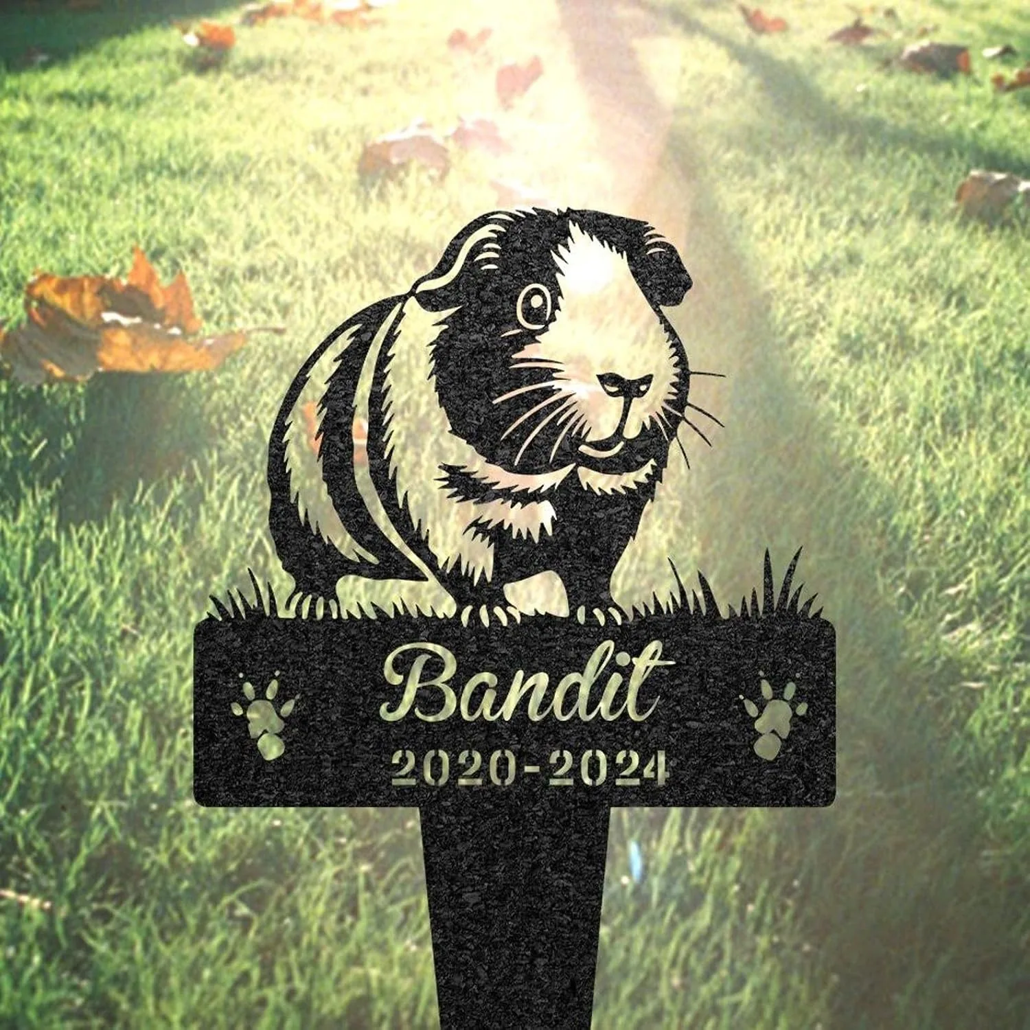 Black metal memorial stake with a guinea pig illustration and the name "Bandit" above dates "2020-2024" placed in sunlit grass