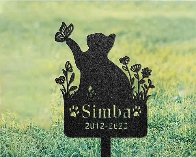 Black metal garden memorial sign shaped like a cat reaching for a butterfly with flowers, engraved “Simba” and date “2012-2022,” staked in grass