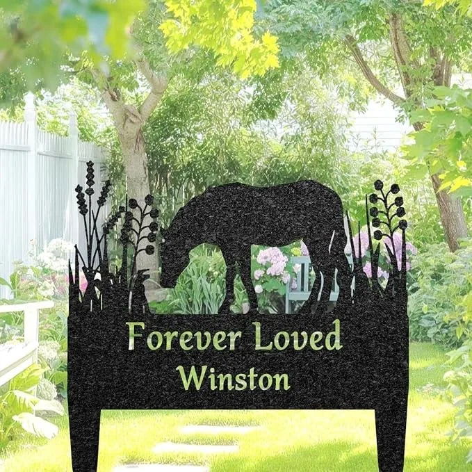 Black garden memorial stake with horse silhouette grazing and plants, green text reading "Forever Loved Winston", placed in a sunny backyard garden