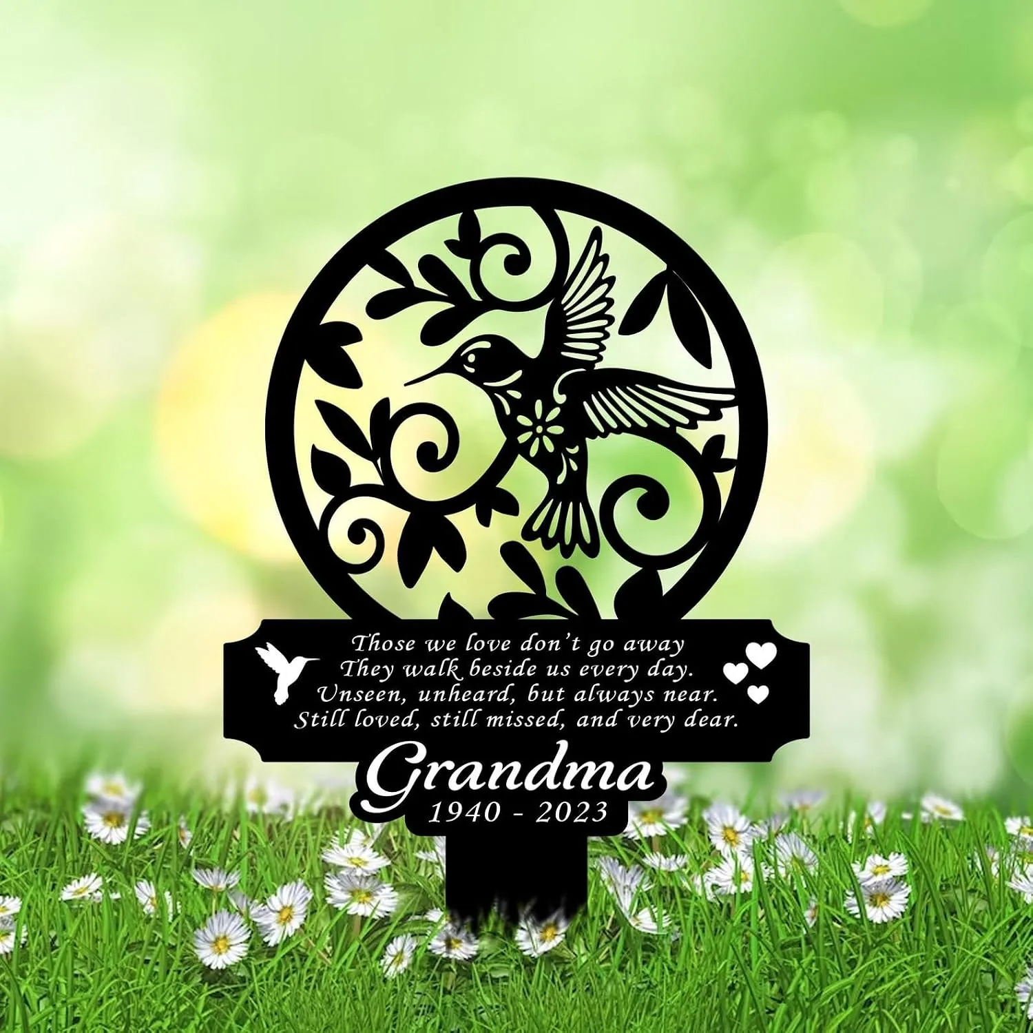 Black circular metal memorial plaque with a hummingbird and leaves, mounted in a daisy-filled lawn; text reads a poem and “Grandma 1940 - 2023”