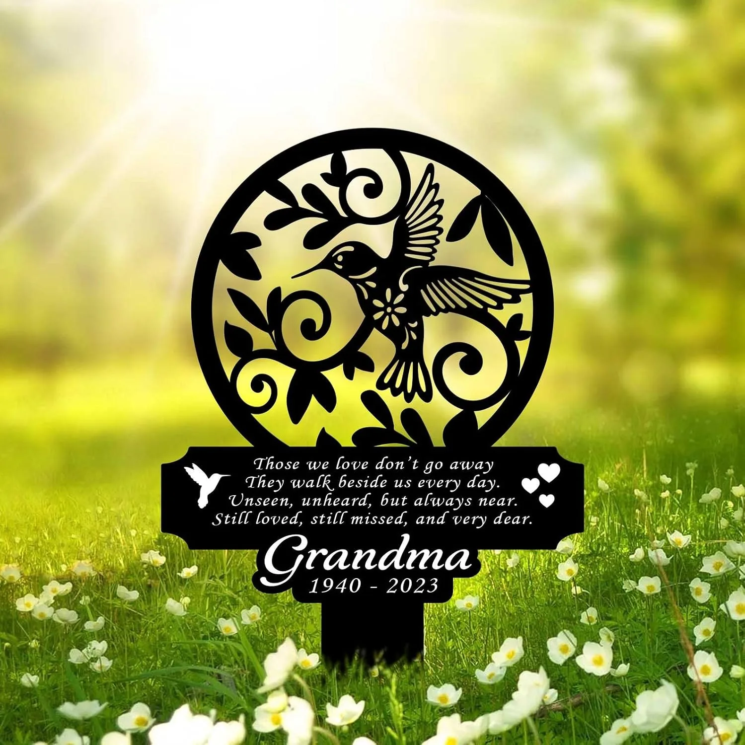 Black metal memorial garden stake with hummingbird and floral cutout, poem text and inscription “Grandma 1940 - 2023” against sunlit grassy field with white flowers