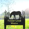 Black metal grave marker silhouette of a grazing horse with floral accents and yellow text reading "Forever Loved Winston" set in sunlit grass
