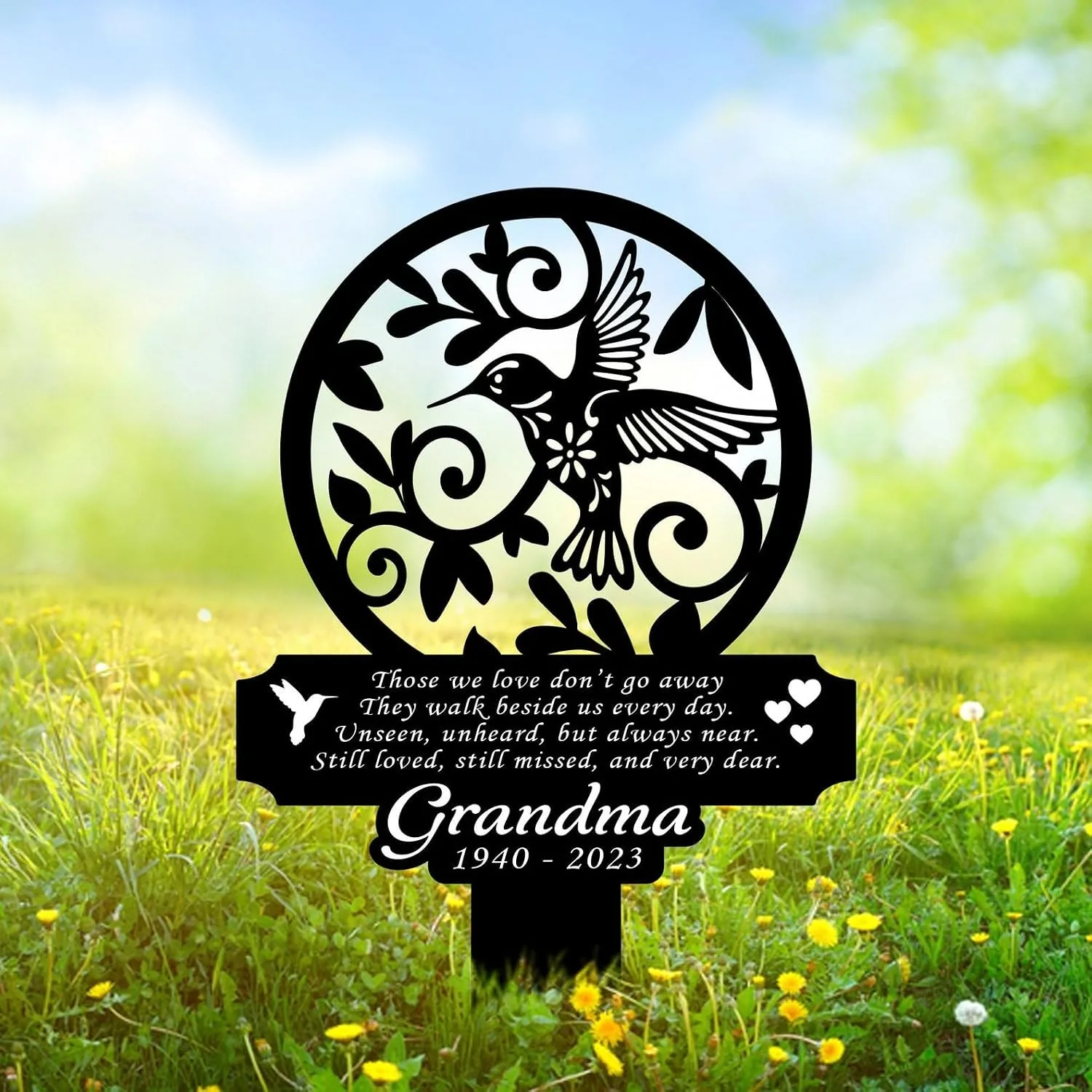 Black metal memorial garden stake with a hummingbird and floral cutout, inscription reading a short poem and 'Grandma 1940 - 2023', placed in grassy field with yellow flowers