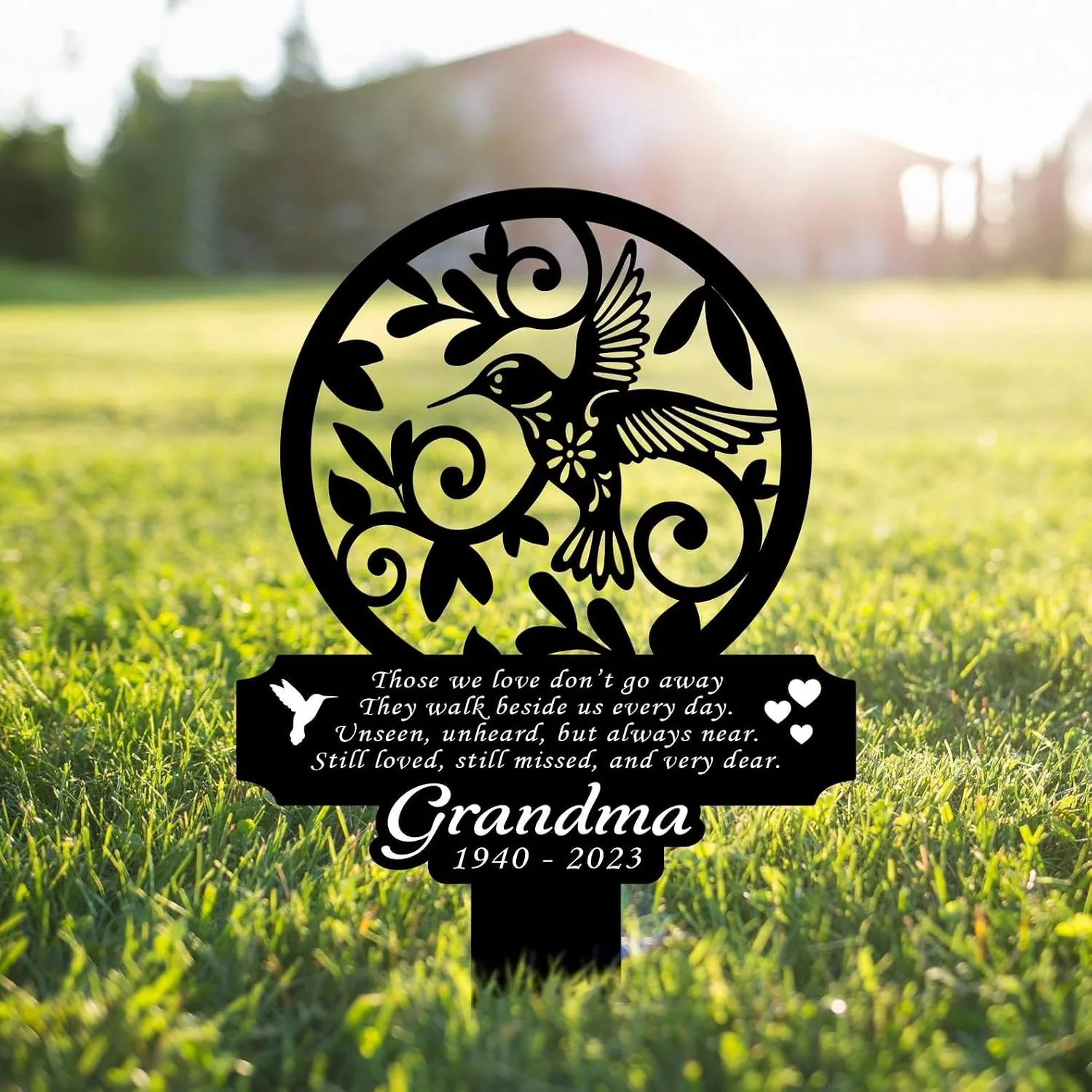 Black metal memorial garden stake in grass with a hummingbird cutout, decorative swirls and text reading “Those we love don’t go away... Grandma 1940 - 2023”
