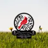 Black metal memorial stake with a red cardinal perched in a circular tree design above engraved text 'When cardinals appear a loved one is near' and name 'LAUREN MILLER 1953 - 2024' set in grassy field with dandelions