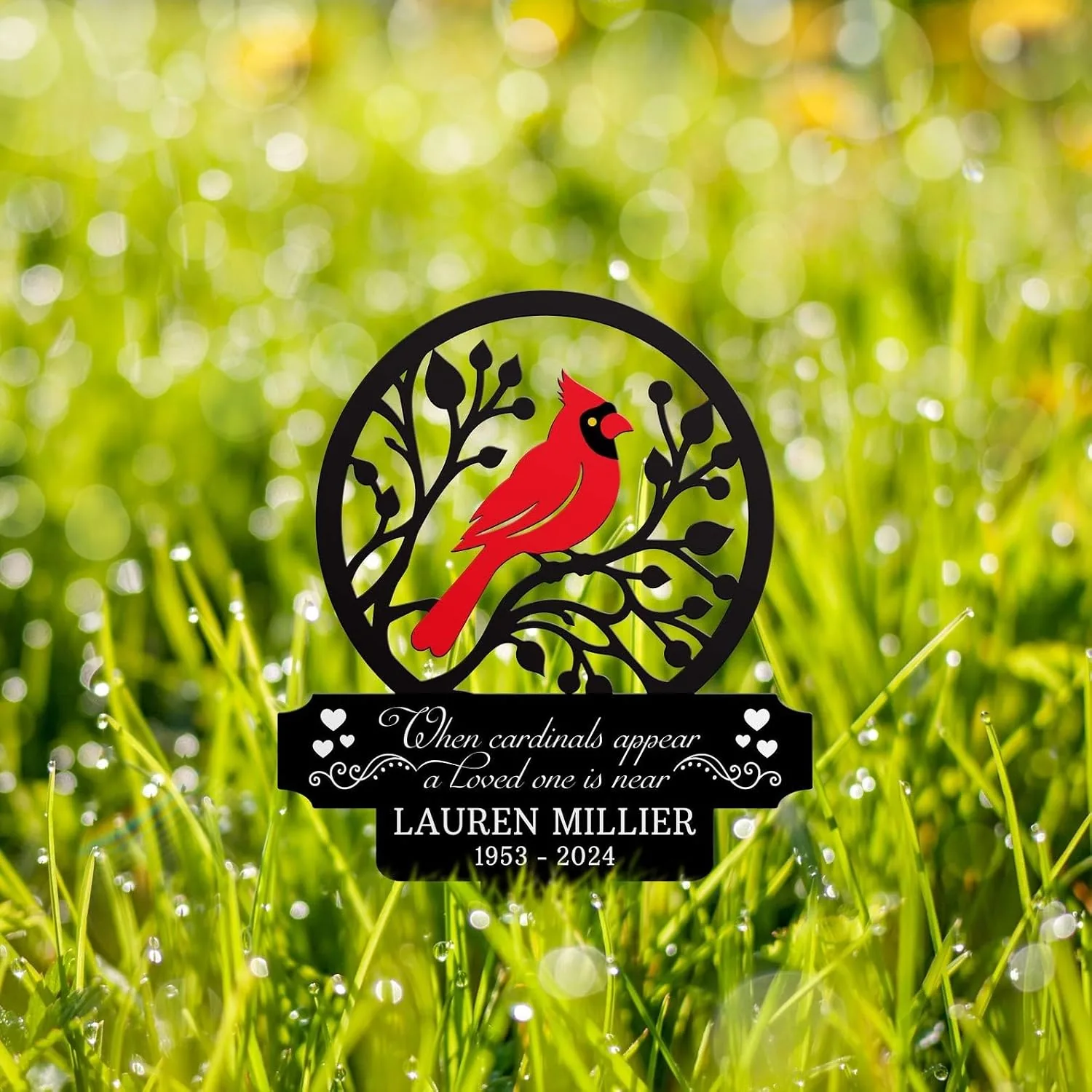 Red cardinal silhouette on a black circular branch memorial sign reading "When cardinals appear a loved one is near" with name Lauren Millier 1953–2024, on dewy green grass background