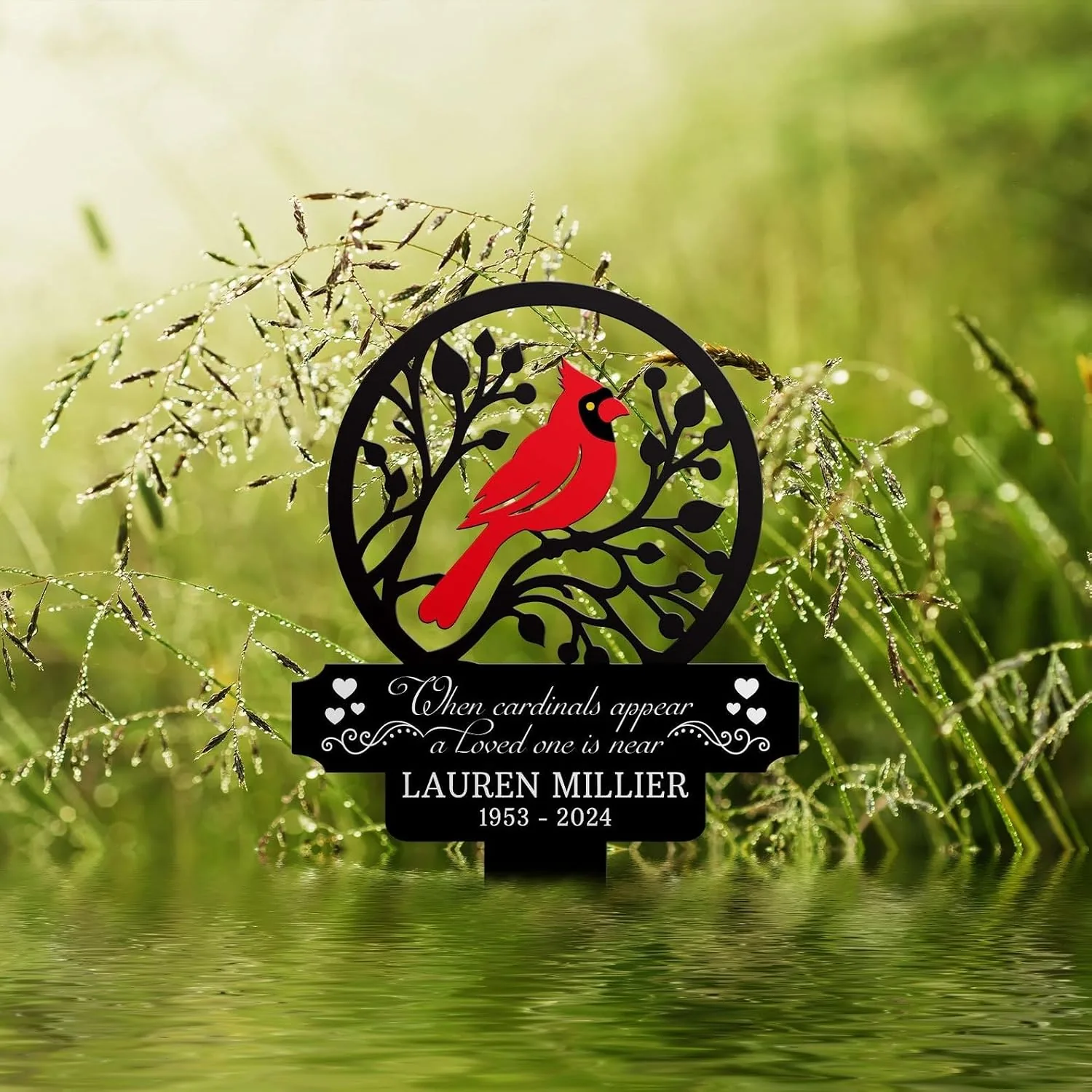 Black circular memorial plaque with a red cardinal perched on branches, text reading "When cardinals appear a Loved one is near" and name "Lauren Millier 1953 - 2024" against a dewy green grass background