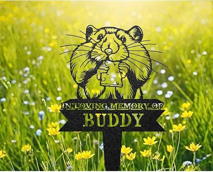 81-1 Black metal memorial stake in grass with cartoon guinea pig holding a heart and text ‘In Loving Memory of Buddy’ among yellow wildflowers