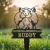83-1 Black metal yard memorial sign of a cartoon hamster holding a square treat with text "In Loving Memory of Buddy" stuck in green grass at sunrise