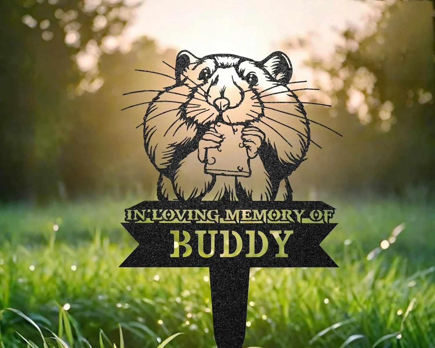 83-1 Black metal yard memorial sign of a cartoon hamster holding a square treat with text "In Loving Memory of Buddy" stuck in green grass at sunrise