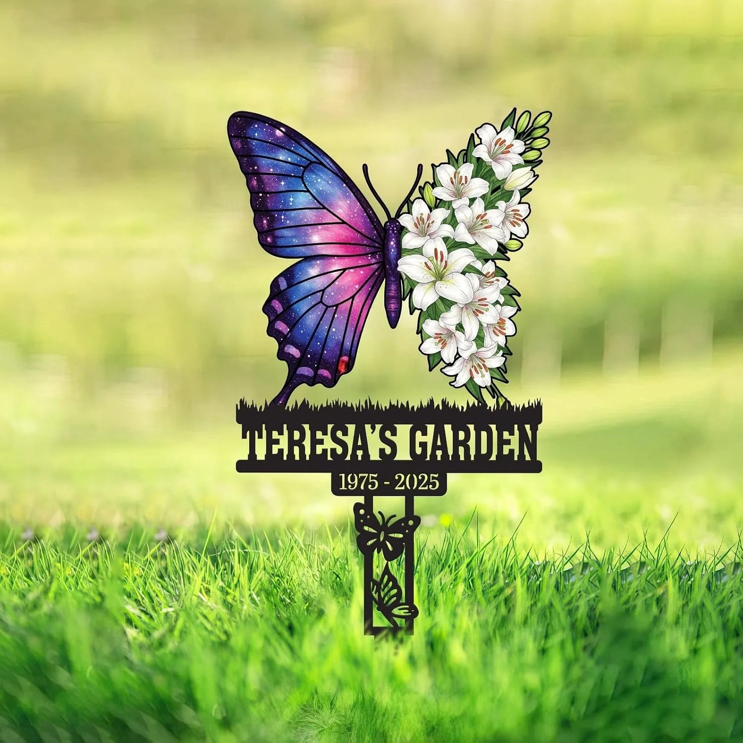Garden memorial sign in grass with a butterfly: left wing galaxy pattern, right wing made of white lilies, black text reading "TERESA'S GARDEN" and years "1975 - 2025"
