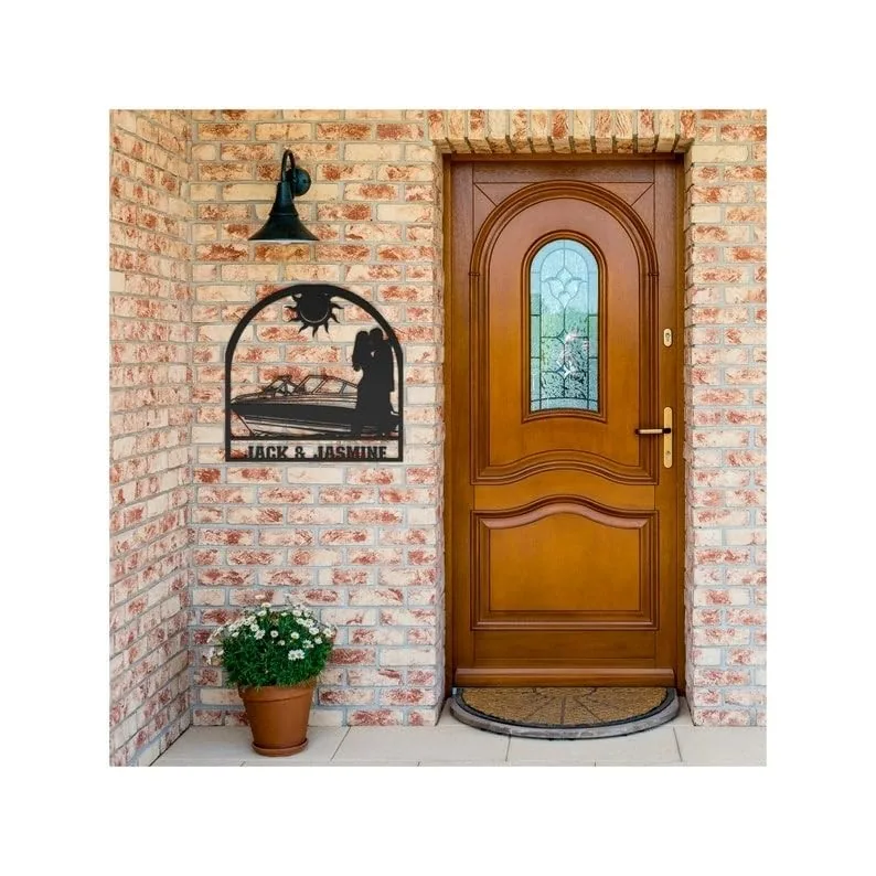 style-16.-Motorboat-Couple-2 Wooden front door with stained glass panel set in a brick entryway, metal wall art reading 'JACK & JASMINE' showing a couple by a boat, potted flowers and black porch light