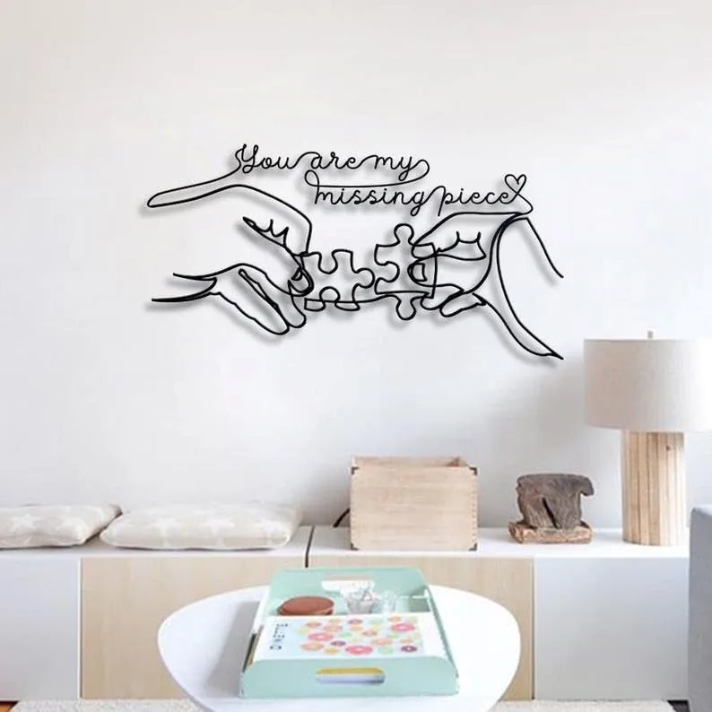Black wire wall art of two hands connecting puzzle pieces with script text “You are my missing piece” above, hung over a modern living room bench and lamp