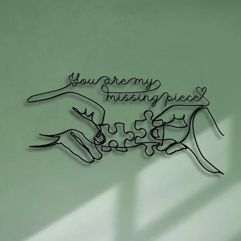 Black wire wall art of two hands connecting puzzle pieces with cursive text reading “You are my missing piece” on a pale green wall
