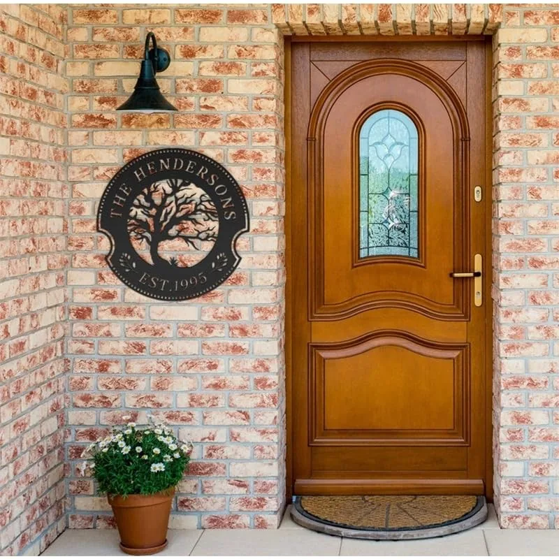 style-19.-Oak-Tree-Couple-3 Wooden front door with decorative glass set in a brick entryway, round metal family name sign reading "The Hendersons EST. 1995" mounted on wall and potted white daisies below