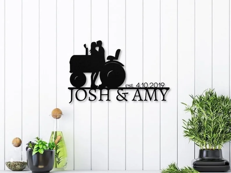 Black metal wall sign of a couple kissing on a tractor above text 'JOSH & AMY' and 'est. 4.10.2019' mounted on white shiplap wall with small potted plants on a shelf