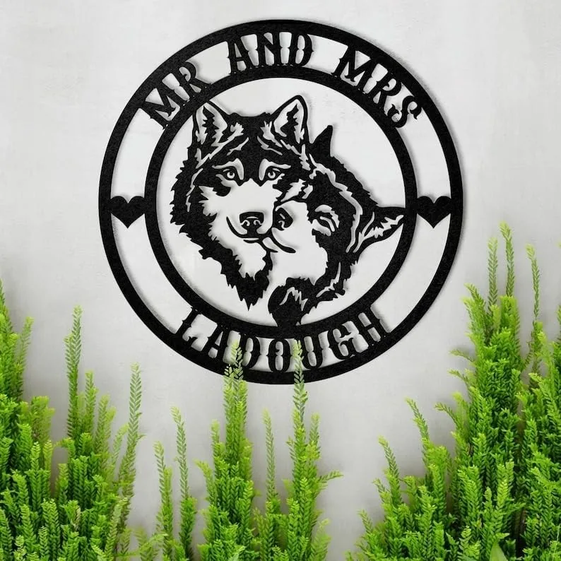style-21.-Wolf-Couple-1-2 Black metal circular wall sign with two wolf heads and text "MR AND MRS" at top and name "LADOUGH" at bottom, displayed on a light wall above green plants