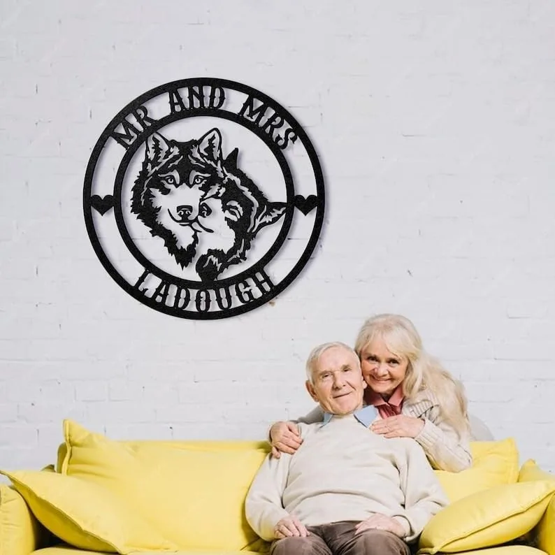 Black circular metal wall art with two wolves and text 'MR AND MRS' and last name 'LADOUGH' hanging on white brick wall above an elderly couple sitting on a yellow couch