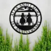 Black round metal wall sign reading "Alex and Laura 1st Anniversary" with silhouette of two cowboys holding hands against a fence, displayed on a light wall above green plants