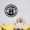Black metal circular wall sign reading "Alfie and Laura 51 Anniversary" with cowboy silhouettes, hung on white brick above an elderly couple on a yellow couch