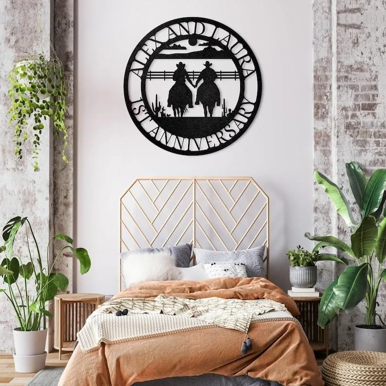 Bedroom with boho decor, rust-colored bedding, plants, and a round black metal wall sign reading ‘Anniversary’ featuring two cowboys on horseback holding hands