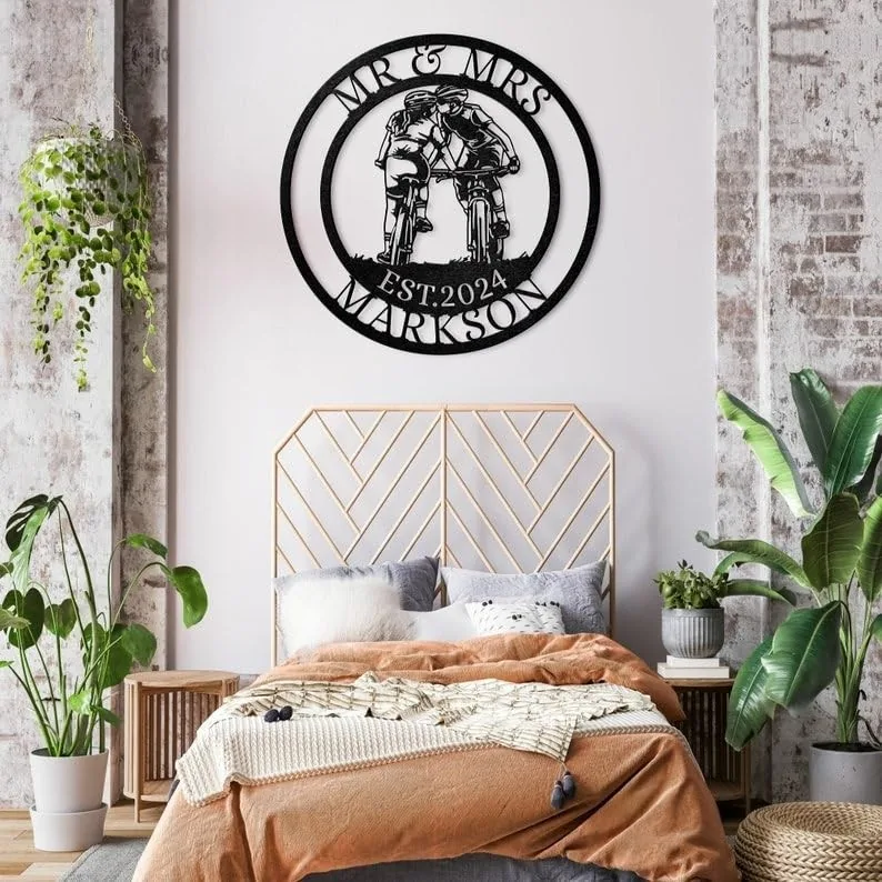 Round black metal wall sign reading "MR & MRS MARKSON EST. 2024" with a kissing couple on bicycles, hanging above a bed in a plant-filled bedroom