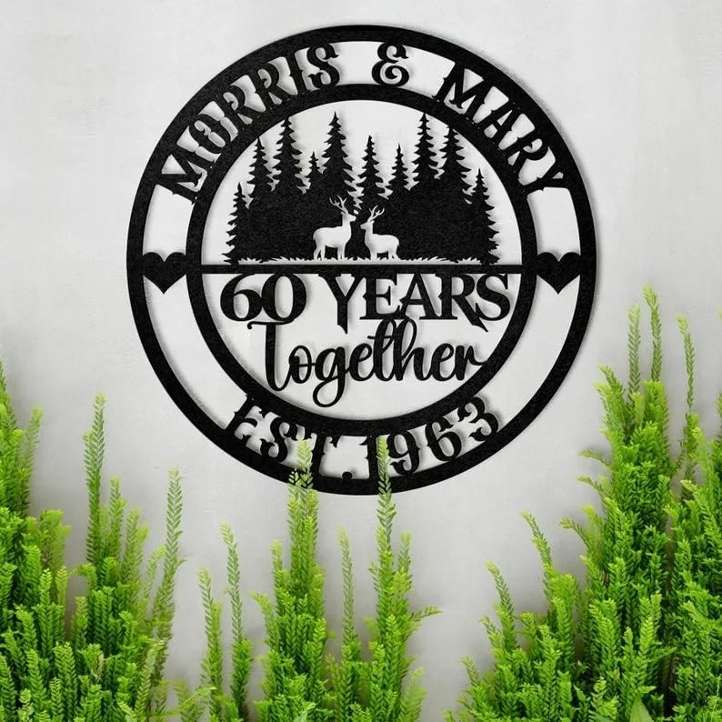 style-25.-Deer-Couple-1 Black circular metal wall sign reading 'Morris & Mary', '60 Years together', 'Est. 1963' with deer and pine tree silhouette, mounted on a light wall above green plants