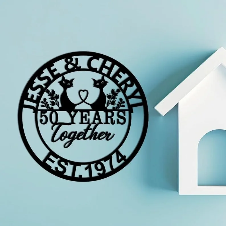 Black circular wall sign reading "JESSE & CHERYL" with two cats forming a heart, "50 YEARS together" and "EST. 1974" mounted on a light blue wall next to a white house-shaped shelf