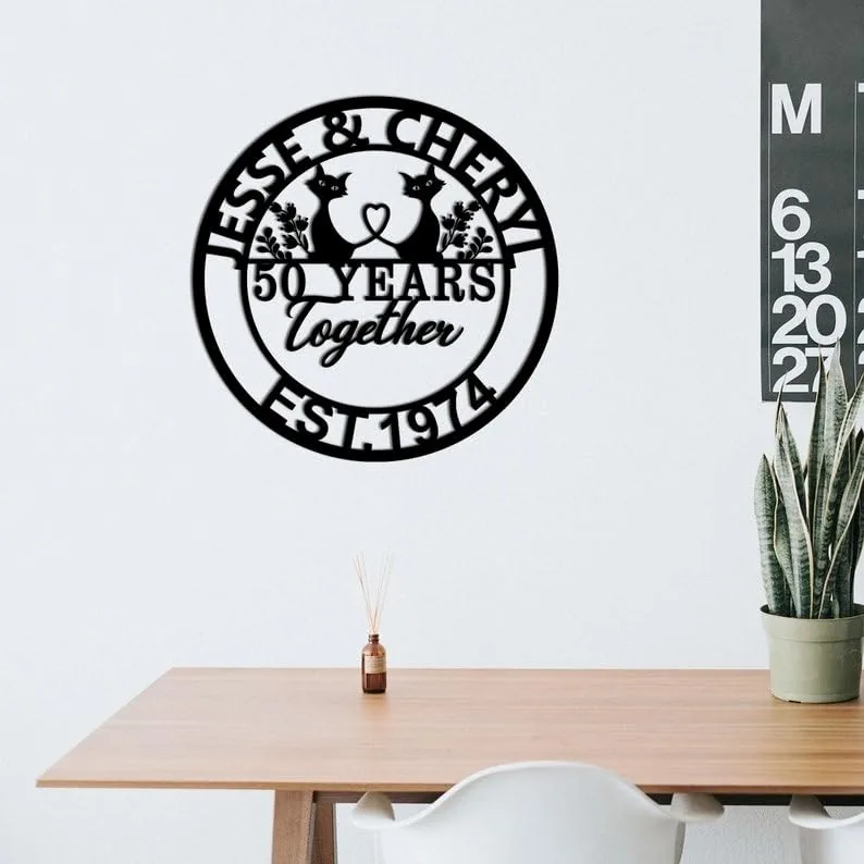 Black circular metal wall sign reading "Jesse & Cheryl 50 Years Together Est. 1974" with two cat silhouettes, displayed above a wooden desk in a minimalist room