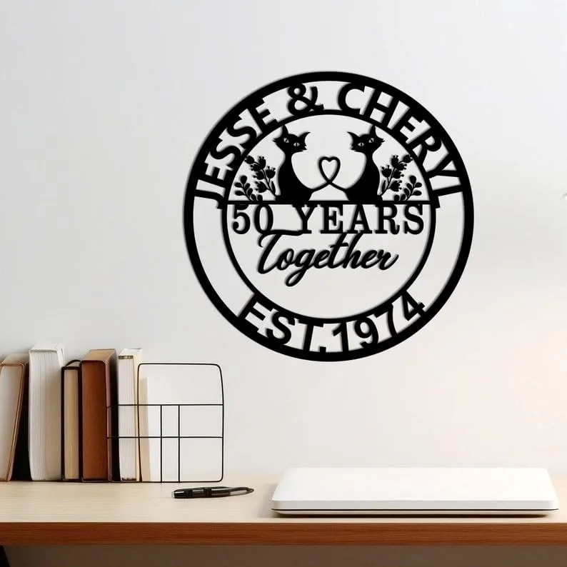 Black circular wall sign reading "Jesse & Cheryl" with two cats, "50 Years Together" and "EST.1974" mounted above a desk with books and laptop