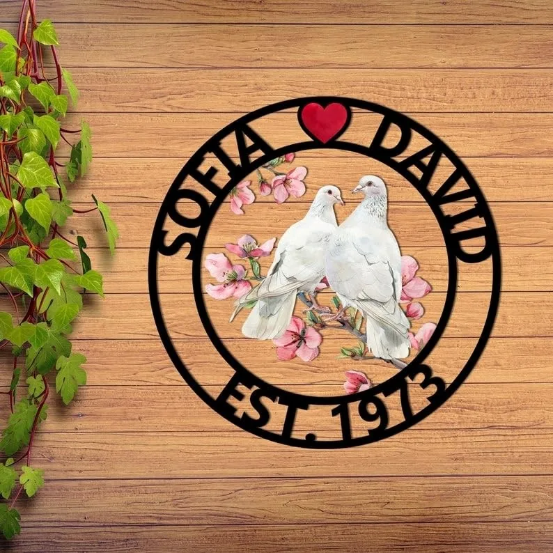 Round wooden wall sign with black cutout ring reading 'SOFIA ♥ DAVID' and 'EST. 1973' surrounding two white doves perched on pink cherry blossoms, displayed on wooden plank background with ivy on left