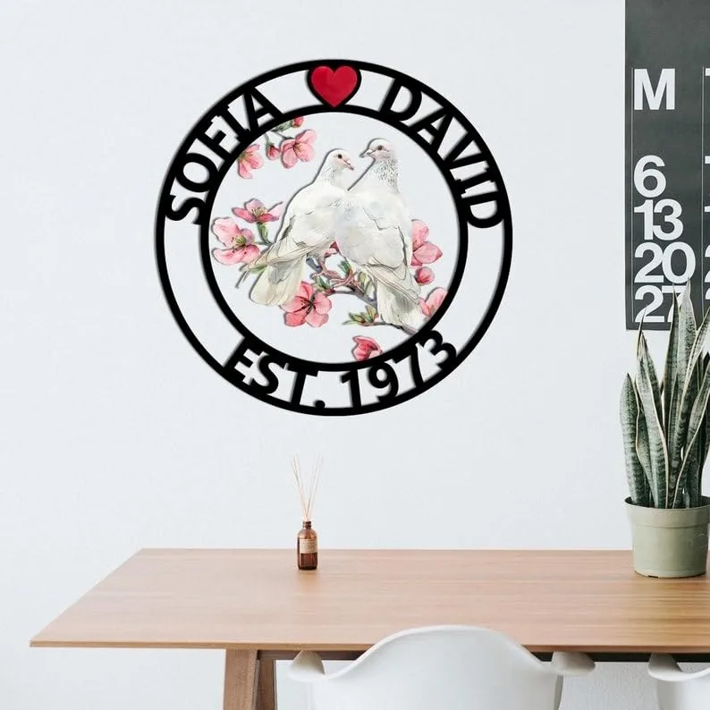 Round black metal wall sign with a red heart at top, names “SOFIA” and “DAVID” around a central illustration of two white doves on pink blossoms and text “EST. 1973,” hanging above a wooden table and plant.