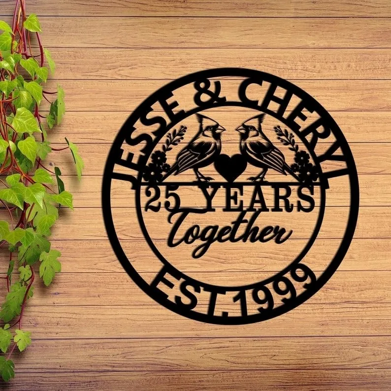 Black circular wooden sign on a wood plank wall reading "Jesse & Cheryl" at top, two cardinals facing a heart with "25 Years Together" in center and "Est. 1999" at bottom
