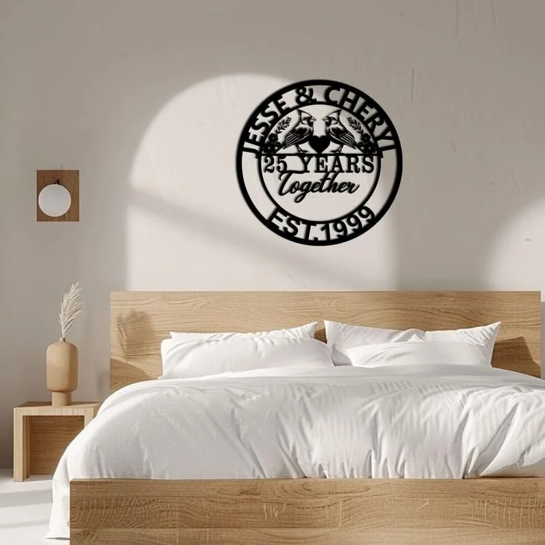Black circular metal wall sign reading "25 Years together" with two birds and "EST 1999" hung above a modern wooden bed in a minimalist bedroom
