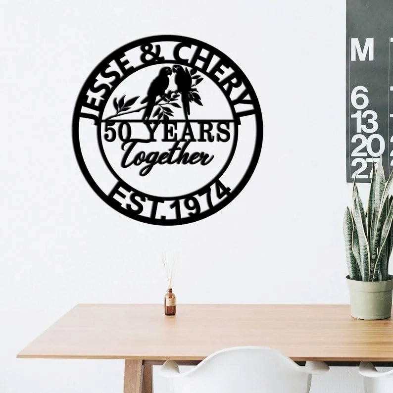 style-29.-Parrot-Couple-3 Black round wall sign reading "Jesse & Cheryl" at top, "50 Years together" in center with two birds on a branch, and "Est. 1974" at bottom, hung above a minimalist desk