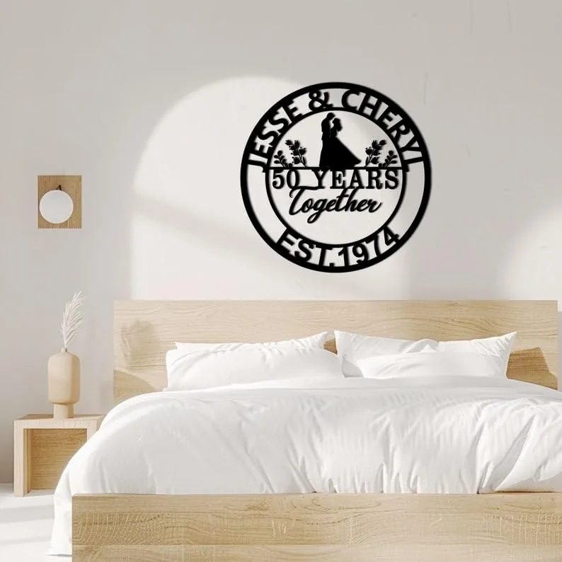 Black round metal wall art above a bed reading 'Jesse & Cheryl 50 Years Together Est. 1974' with silhouette couple and floral accents in a minimalist bedroom