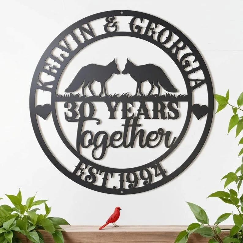 Black circular metal wall sign reading "KEVIN & GEORGIA" at top, two fox silhouettes, "30 YEARS together" in center and "EST. 1994" at bottom, mounted on a white wall above a shelf with plants and a small red bird figurine.