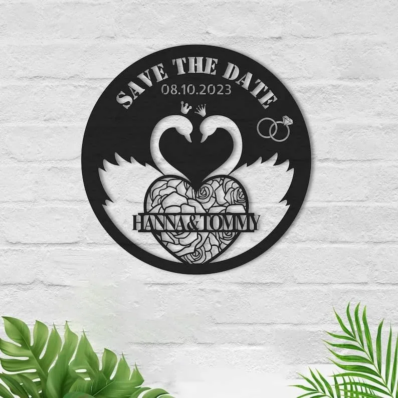 Round black metal wall art with two swans forming a heart, floral heart center reading 'HANNA & TOMMY', 'SAVE THE DATE' and date 08.10.2023, mounted on white brick wall