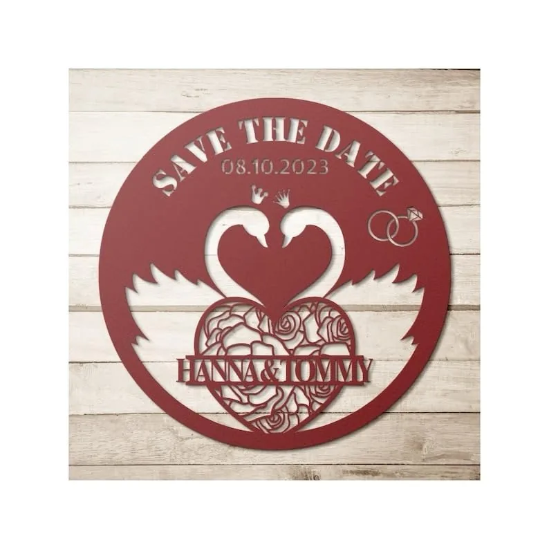 Round red wooden wedding sign on a whitewashed plank background reading "SAVE THE DATE" with date 08.10.2023, two swans forming a heart, floral heart and names "HANNA & TOMMY"