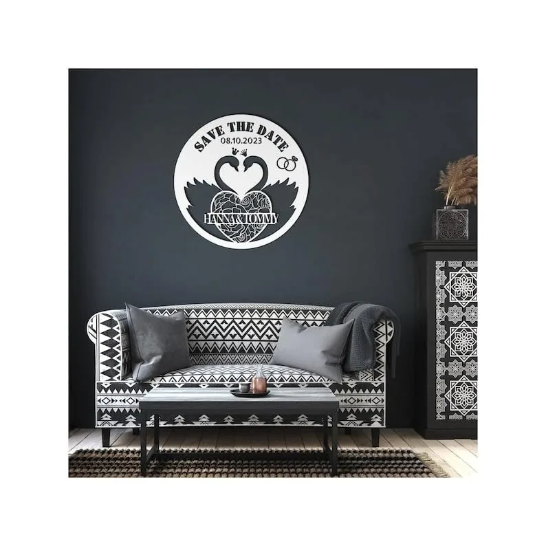 Round white wooden 'Save the Date' wall sign with two swans forming a heart, wedding rings, date 08.10.2023 and names over a dark gray living room wall above patterned black-and-white sofa