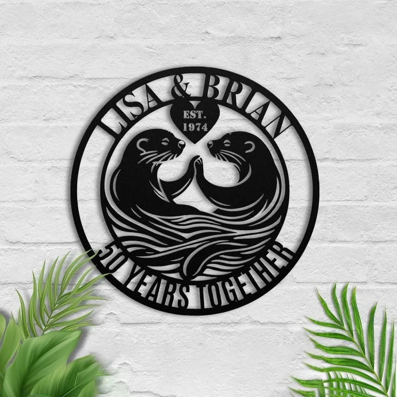 style-35.-Otter-Anniversary-1 Black metal wall art of two otters touching paws inside a circle reading "LISA & BRIAN" with a heart saying "EST. 1974" and "50 YEARS TOGETHER" on a white brick wall with green leaves