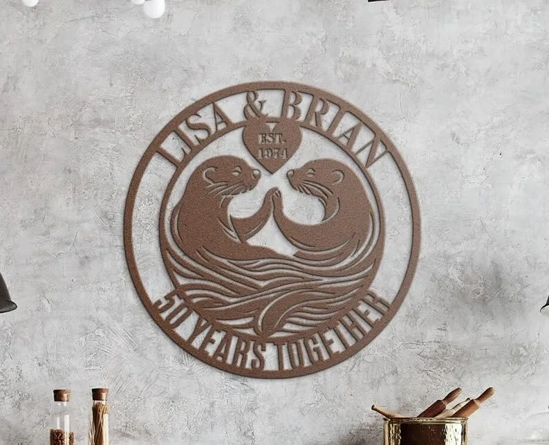 style-35.-Otter-Anniversary-3 Round metal wall sign with two otters facing each other, heart at top reading "EST. 1971", names "Lisa & Brian" along top and "50 Years Together" along bottom, hung on a textured gray wall above kitchen counter