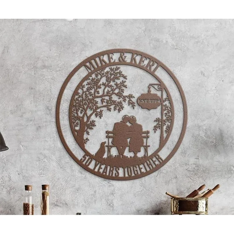 Rust-colored metal wall art showing a couple sitting on a bench under a tree with a sign reading 'EST 1974', names 'MIKE & KERI' at top and '50 YEARS TOGETHER' along bottom, mounted on a textured gray wall